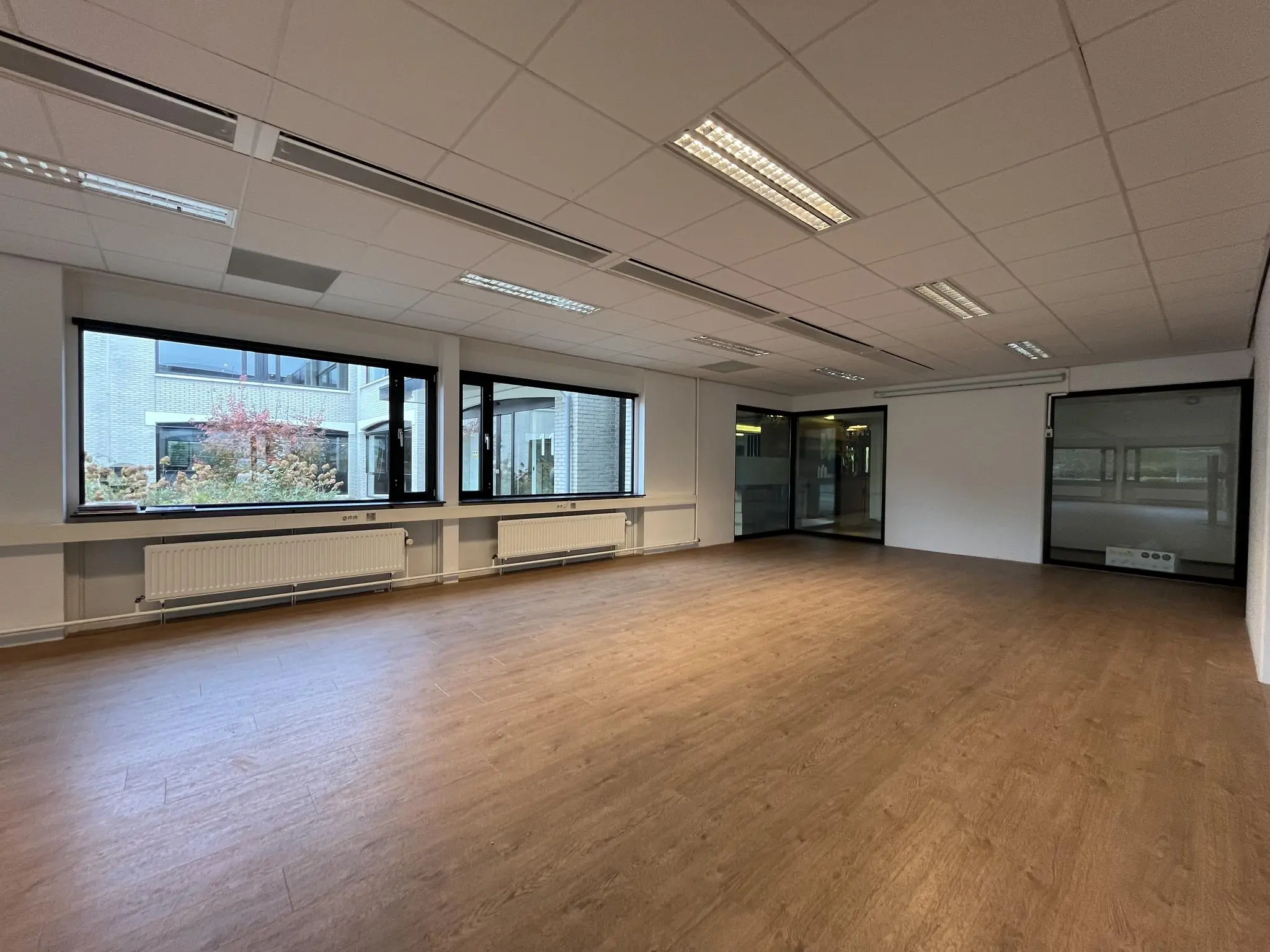 Leeg kantoorruimte aan de Burgemeester Verderlaan met grote ramen, houten vloer en systeemplafond.